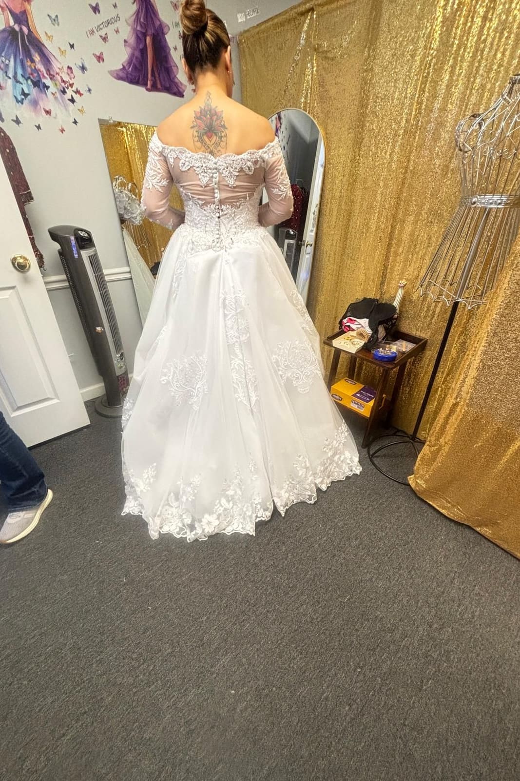 A bride at her fitting, seen from behind in an off-the-shoulder lace wedding gown