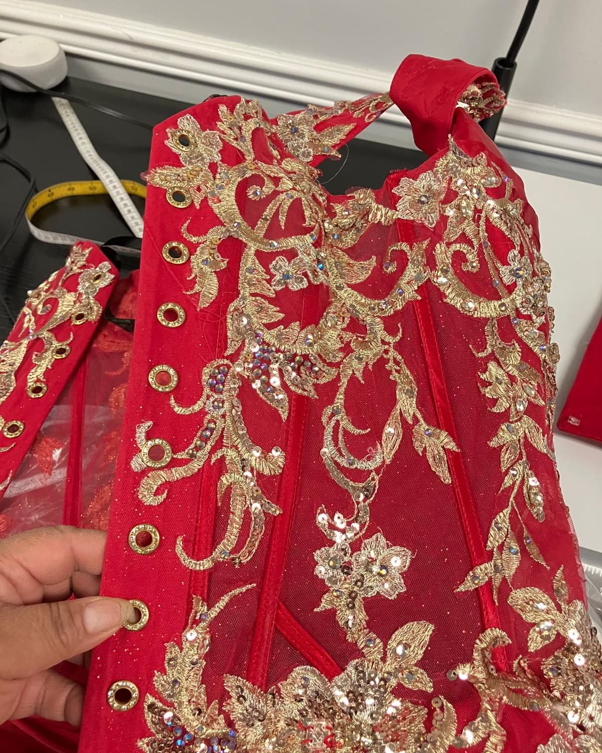 A hand holding the red corset of a quinceañera gown, embroidered in gold with sequins and eyelets