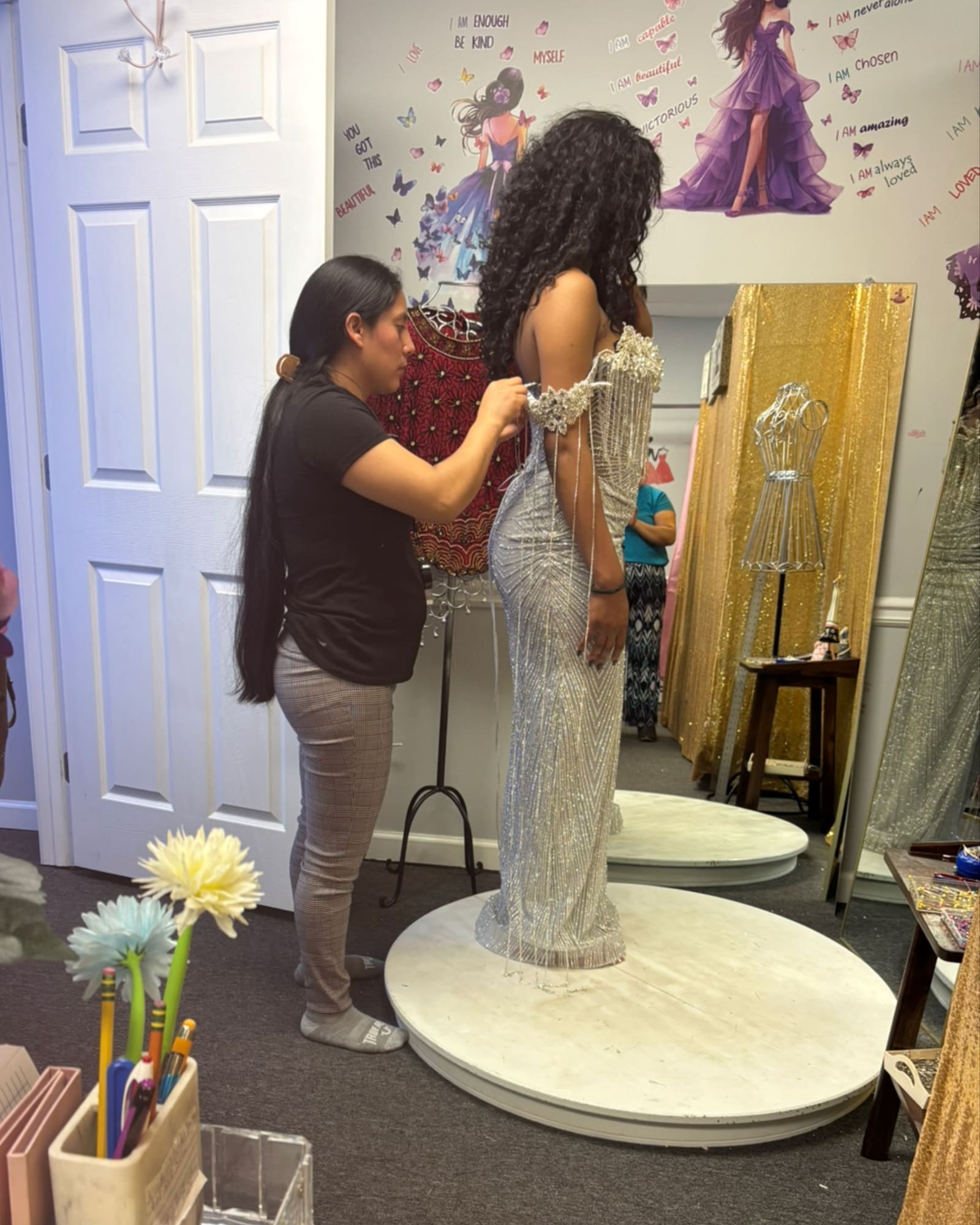 Yolanda pinning a silver beaded gown on a client standing on a fitting platform
